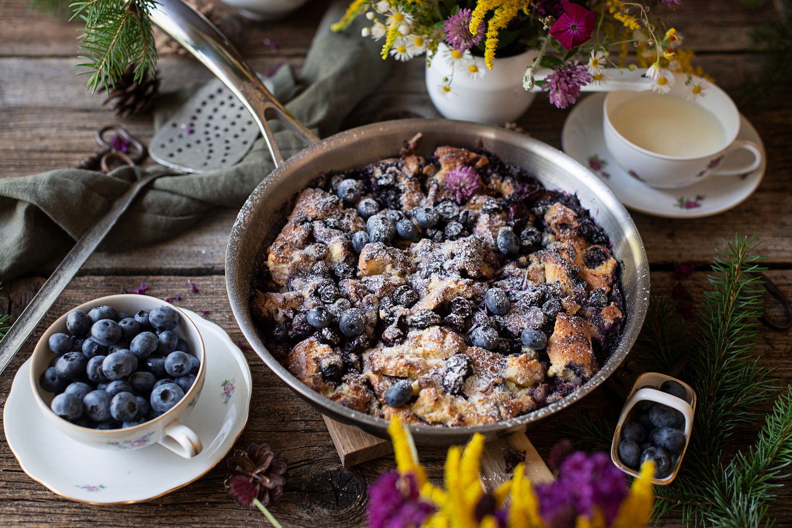 Bratpfanne mit Heidelbeerauflauf, von oben fotografiert, umgeben von Blumen und Geschirr.