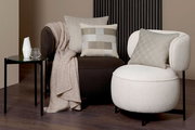 Zwei moderne Sessel mit dekorativen Kissen in einem stilvollen Wohnzimmer. Der linke Sessel ist dunkelbraun mit einem beigen Strickplaid und einem karierten Kissen. Der rechte Sessel ist cremefarben mit einem gemusterten Kissen. Perspektive: Frontalansich