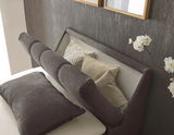 Boxspringbett mit gepolstertem Kopfteil, seitlicher Blickwinkel, dekoriert mit Kissen und Blumen.