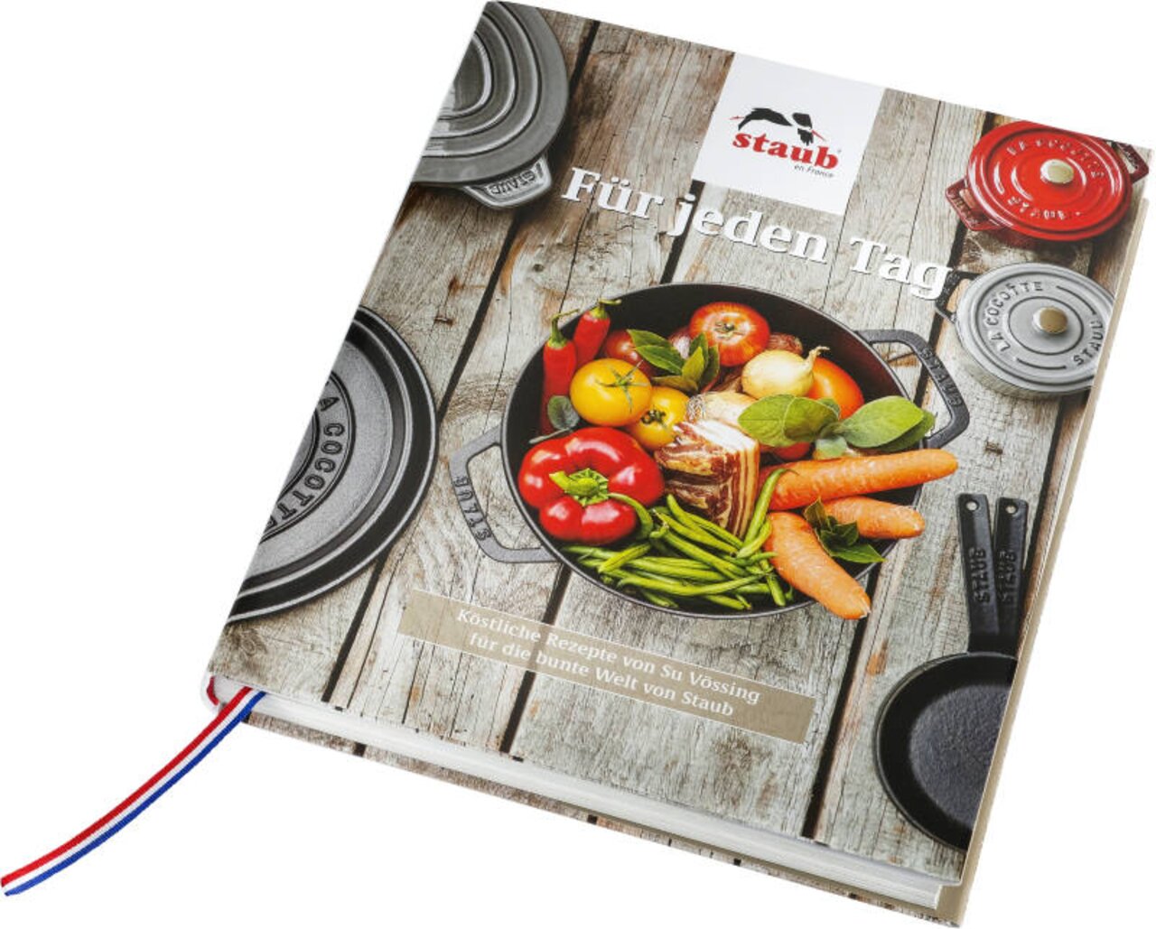Het STAUB-kookboek met de titel 'Für jeden Tag', afgebeeld vanuit een schuin perspectief, toont een pan met verse groenten op een houten tafel.