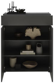 Schwarze Kommode mit geöffneter Tür, dekoriert mit Kerzenhaltern und Schalen auf der Oberseite, und verschiedenen Dekorationsobjekten sowie Büchern in den Regalfächern, frontal betrachtet.