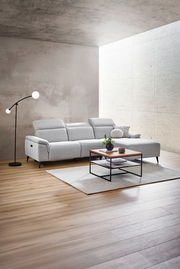 Moderne Longchairkombination in einem hellen Wohnzimmer, seitliche Perspektive. Die graue Couch steht auf einem Holzboden, daneben eine Stehlampe und ein Couchtisch mit Dekoration.