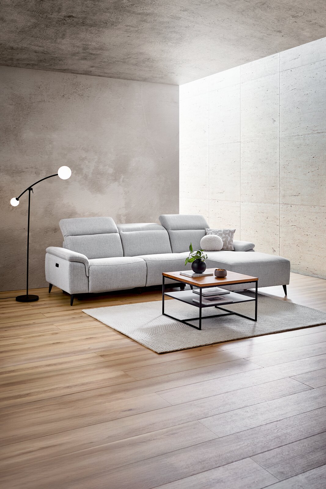 Moderne Longchairkombination in einem hellen Wohnzimmer, seitliche Perspektive. Die graue Couch steht auf einem Holzboden, daneben eine Stehlampe und ein Couchtisch mit Dekoration.