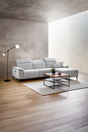 Moderne Longchairkombination in einem hellen Wohnzimmer, seitliche Perspektive. Die graue Couch steht auf einem Holzboden, daneben eine Stehlampe und ein Couchtisch mit Dekoration.