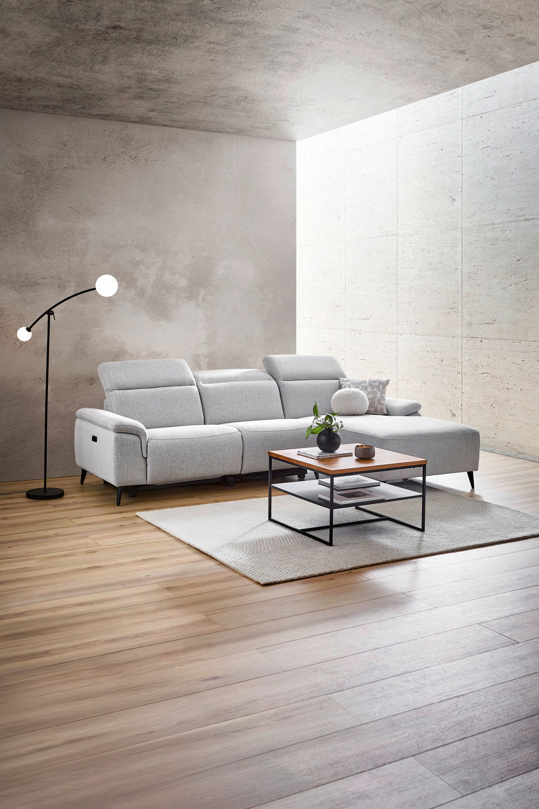 Moderne Longchairkombination in einem hellen Wohnzimmer, seitliche Perspektive. Die graue Couch steht auf einem Holzboden, daneben eine Stehlampe und ein Couchtisch mit Dekoration.