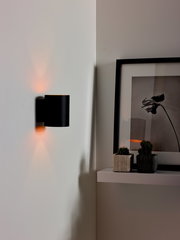 Moderne schwarze Wandleuchte XERA von Lucide, seitliche Perspektive, beleuchtet eine weiße Wand mit warmem Licht.