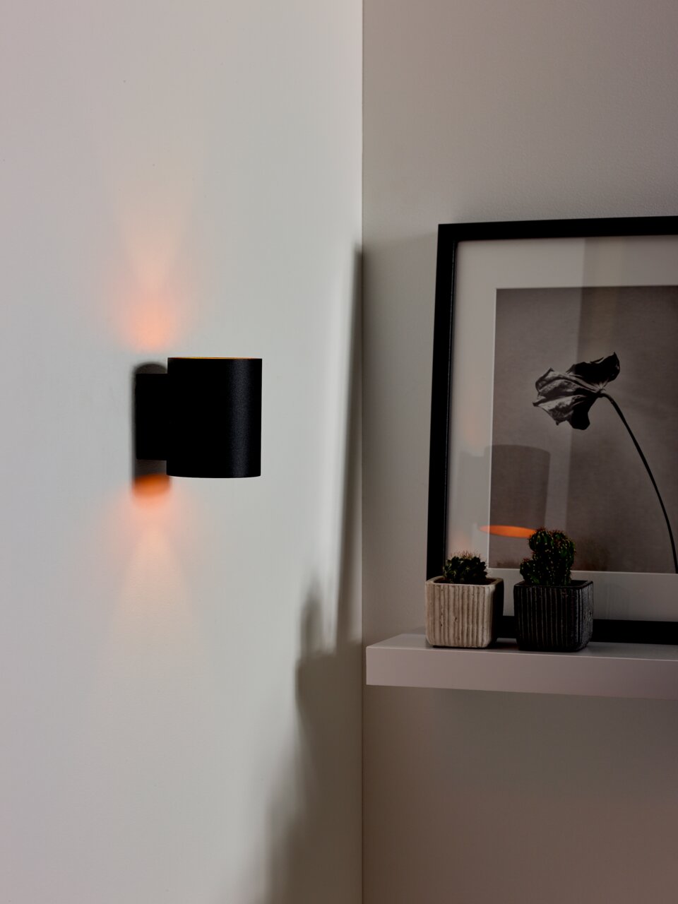 Moderne schwarze Wandleuchte XERA von Lucide, seitliche Perspektive, beleuchtet eine weiße Wand mit warmem Licht.
