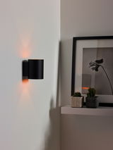 Moderne schwarze Wandleuchte XERA von Lucide, seitliche Perspektive, beleuchtet eine weiße Wand mit warmem Licht.