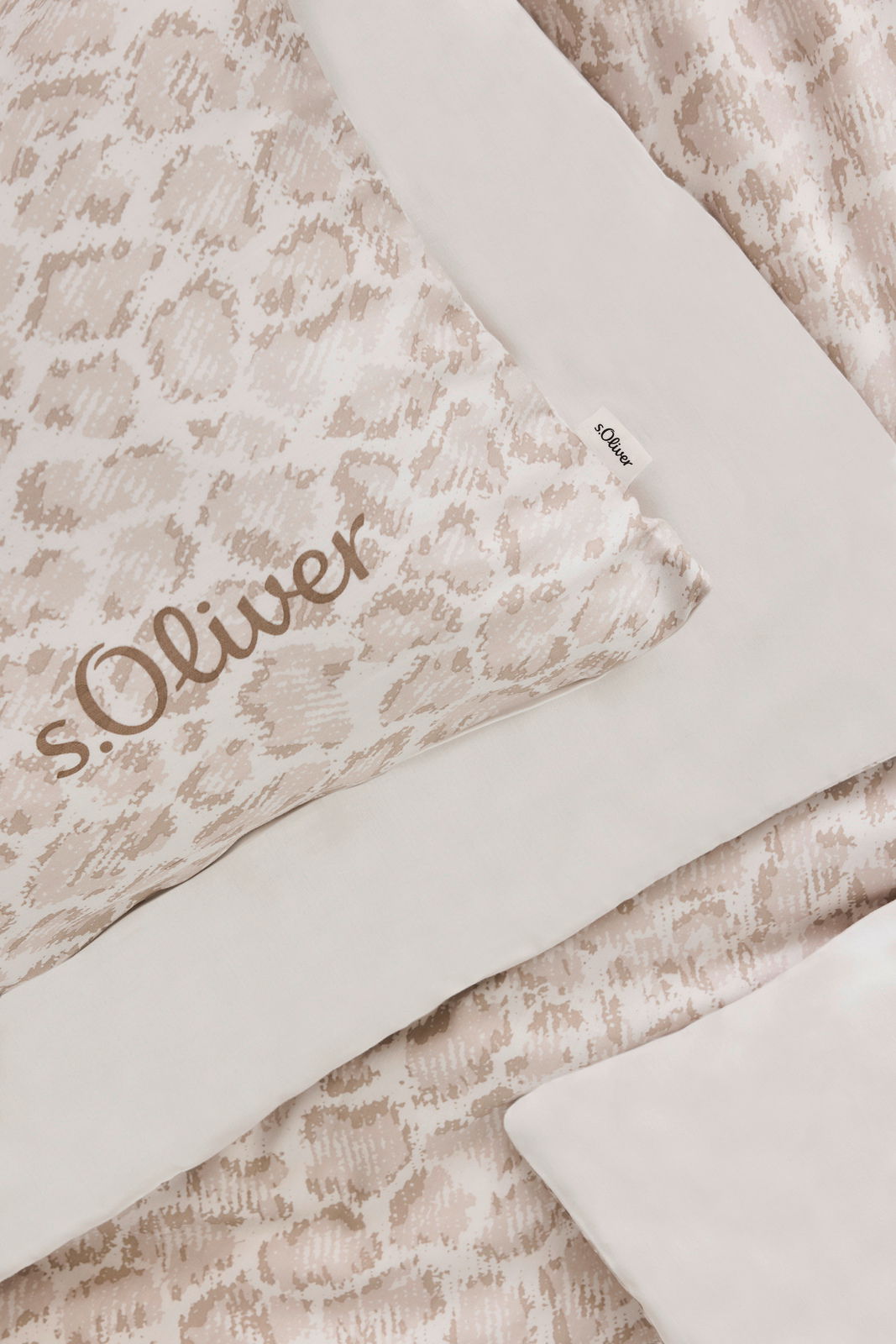 Nahaufnahme einer s.Oliver Satinbettwäsche in Beige mit dezentem Muster und Markenlogo, von oben fotografiert.