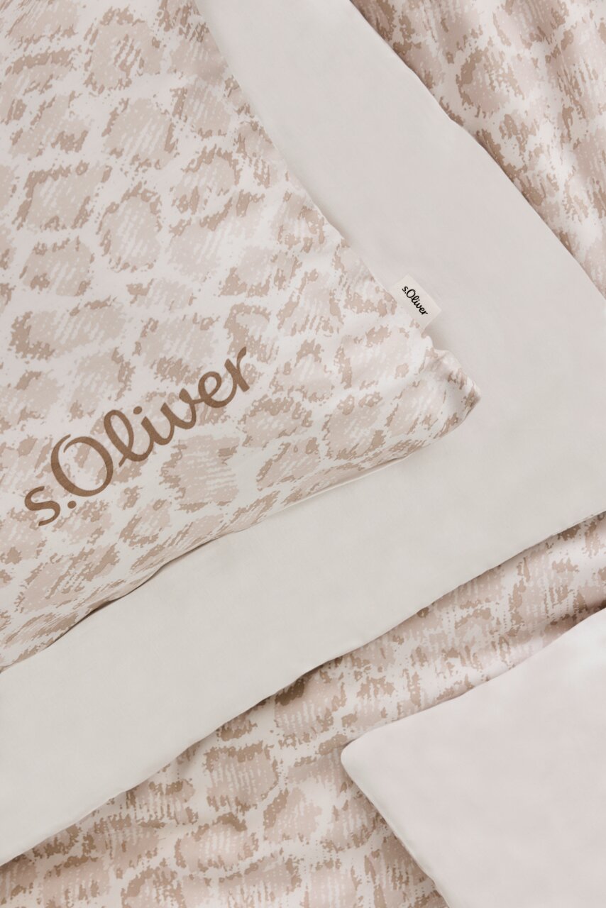 Nahaufnahme einer s.Oliver Satinbettwäsche in Beige mit dezentem Muster und Markenlogo, von oben fotografiert.