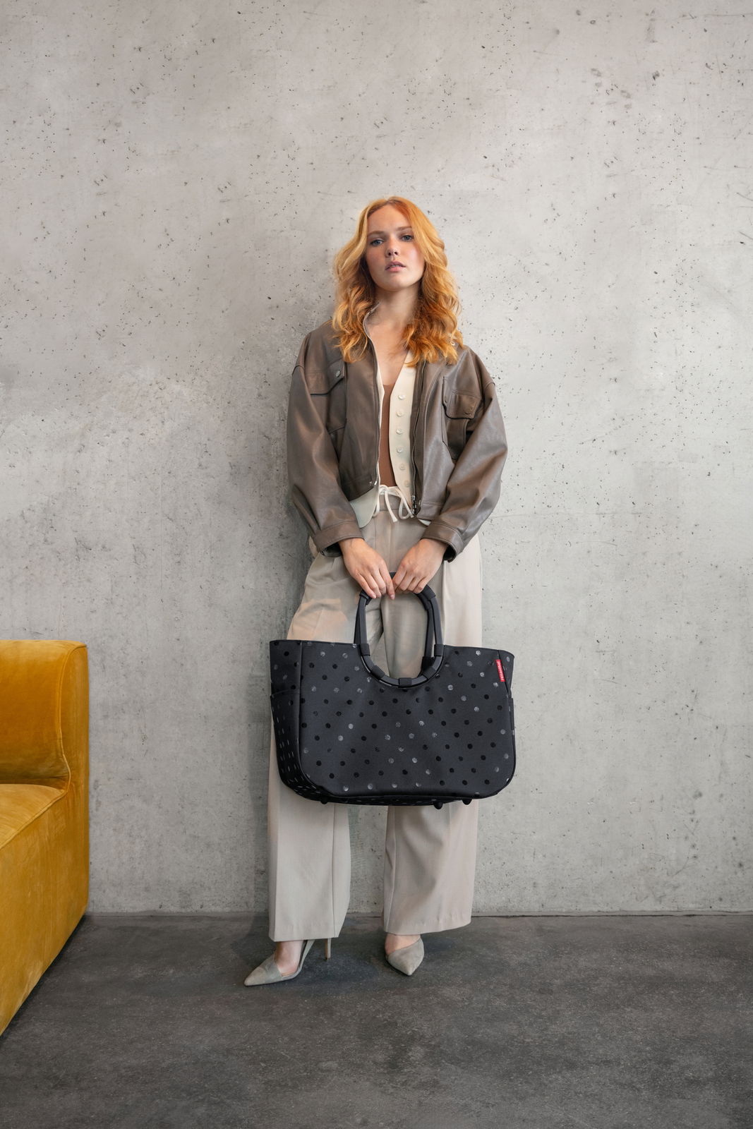 Frau steht vor einer Betonwand und hält eine große schwarze Tasche mit glänzenden Punkten in der Hand, frontal zur Kamera.