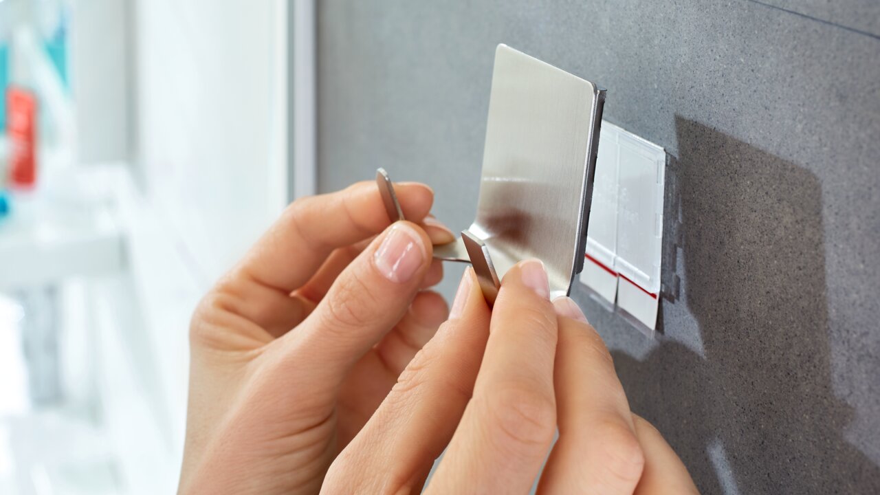 Nahaufnahme von Händen, die einen tesa Powerstrip Waterproof an einer Wand anbringen. Die Perspektive zeigt die Hände von vorne, während sie den Klebestreifen befestigen.