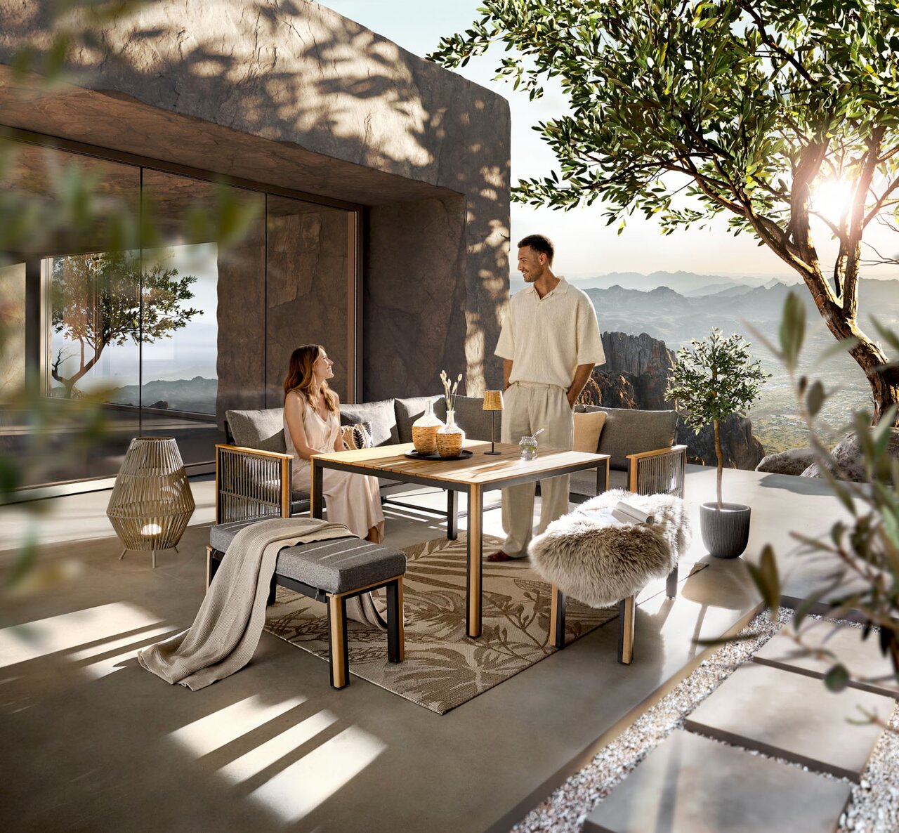 Moderne Outdoor-Lounge auf einer Terrasse mit Holz-Esstisch, gepolsterter Eckbank und Sesseln, Dekoration und Teppich im warmen Sonnenlicht mit Bergpanorama im Hintergrund, aufgenommen aus leicht erhöhter Frontperspektive