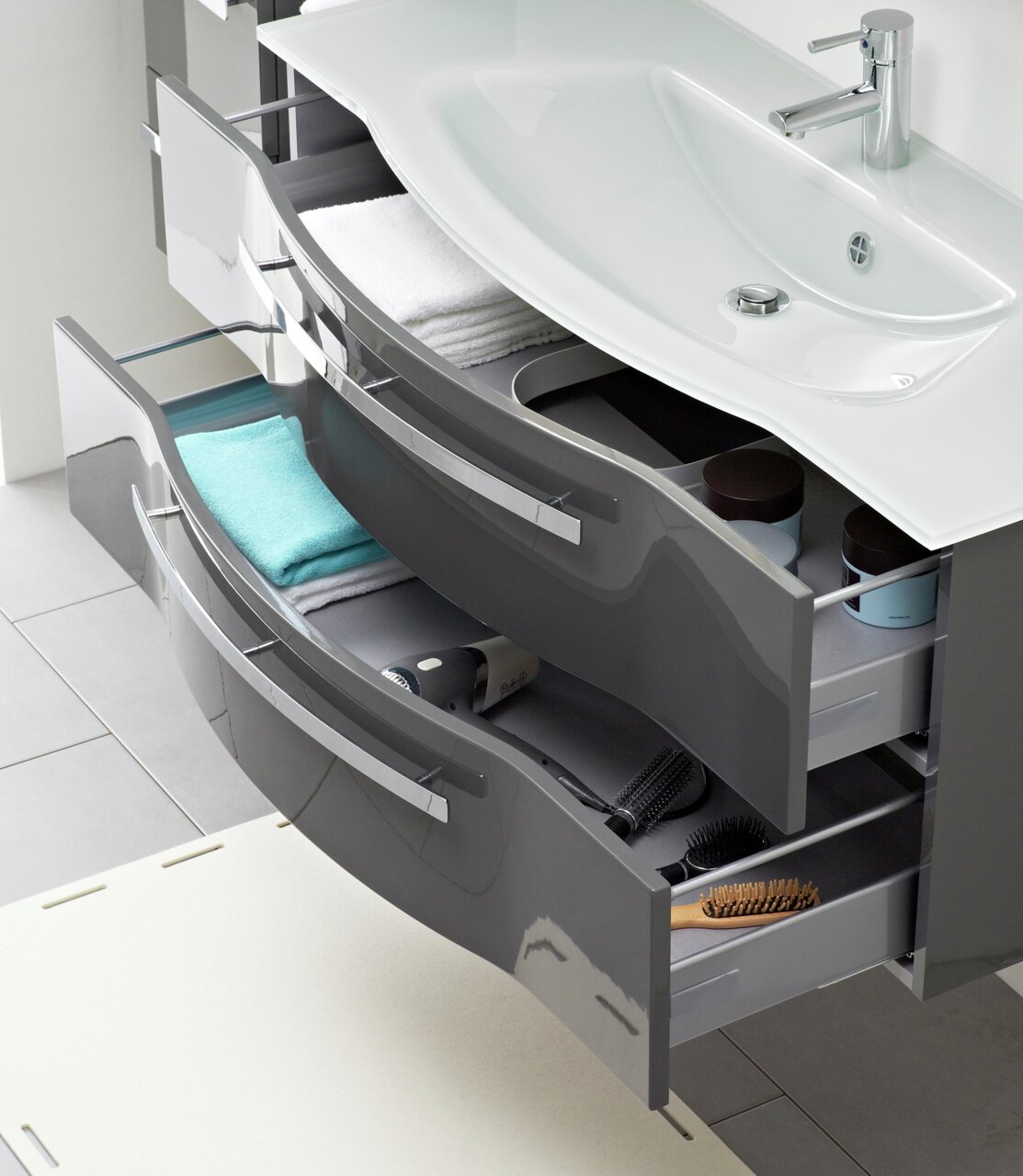 Moderner Badblock mit geschwungenem Waschbecken und zwei offenen Schubladen, gefüllt mit Handtüchern und Pflegeprodukten, aus der Vogelperspektive.
