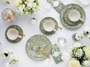Draufsicht auf eine elegant gedeckte Tafel mit grünem, floral gemustertem Geschirr (Teller und Tassen), goldfarbenem Besteck, Gläsern, Servietten und weißen Blumenarrangements auf weißem Hintergrund.