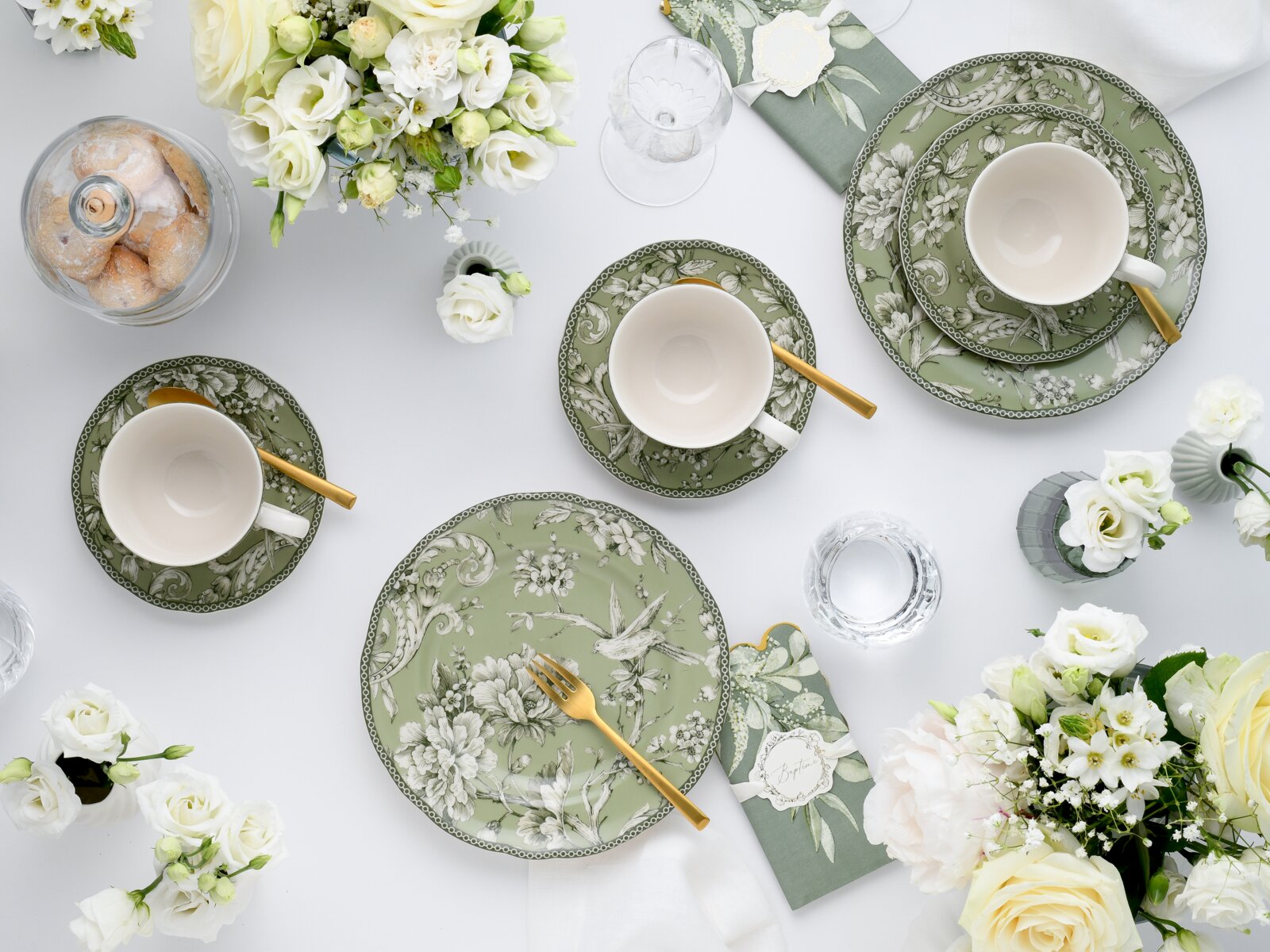Draufsicht auf eine elegant gedeckte Tafel mit grünem, floral gemustertem Geschirr (Teller und Tassen), goldfarbenem Besteck, Gläsern, Servietten und weißen Blumenarrangements auf weißem Hintergrund.