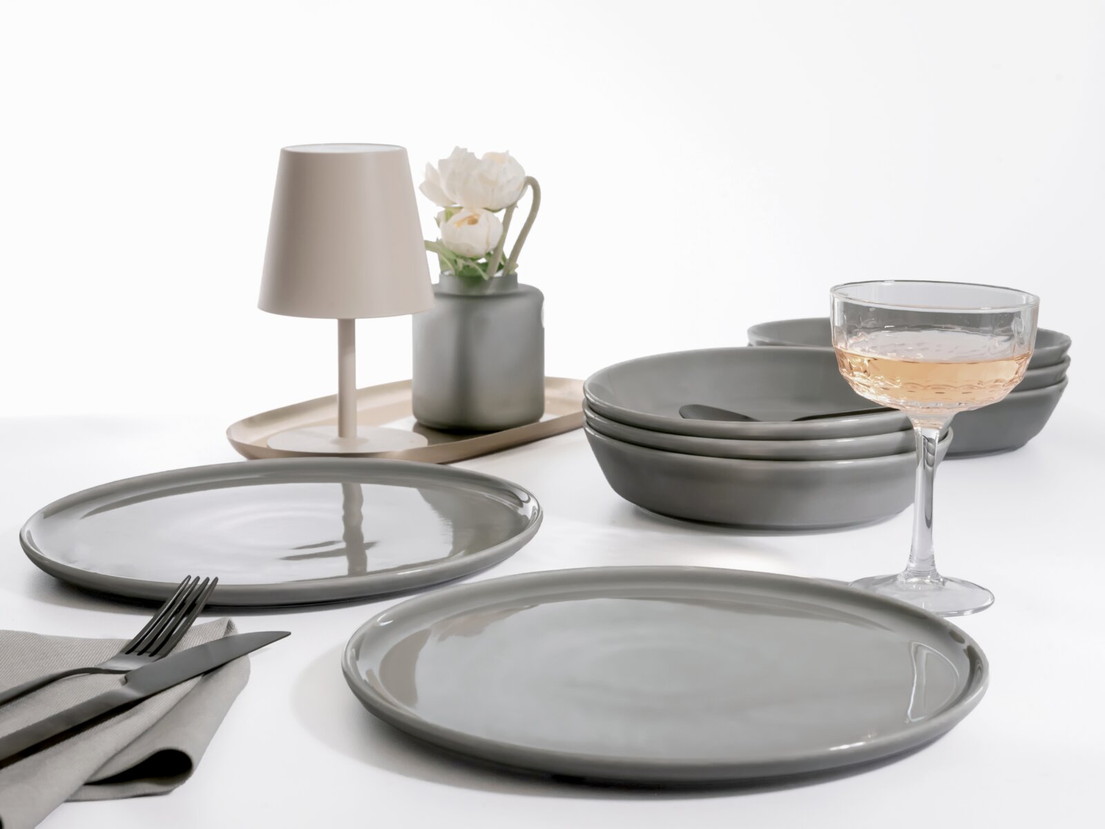 Modernes Tischgedeck mit grauen Keramiktellern und -schalen, Weinglas mit Rosé, kleiner Tischlampe und Vase auf Tablett, aufgenommen aus leicht erhöhter schräger Perspektive (3/4-Ansicht) vor weißem Hintergrund.