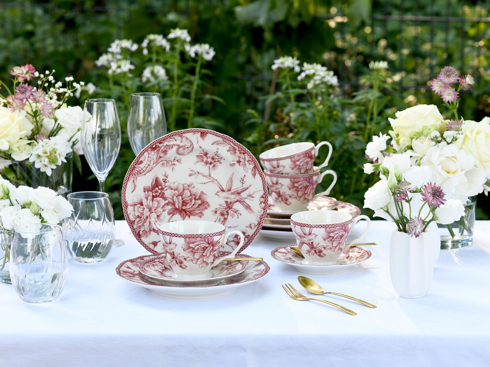 Romantisches Porzellan-Kaffeeservice in Weiß mit bordeauxrotem floralen Muster, Tassen und Teller auf weiß gedecktem Tisch im Garten, frontal aus leicht erhöhter Perspektive fotografiert.