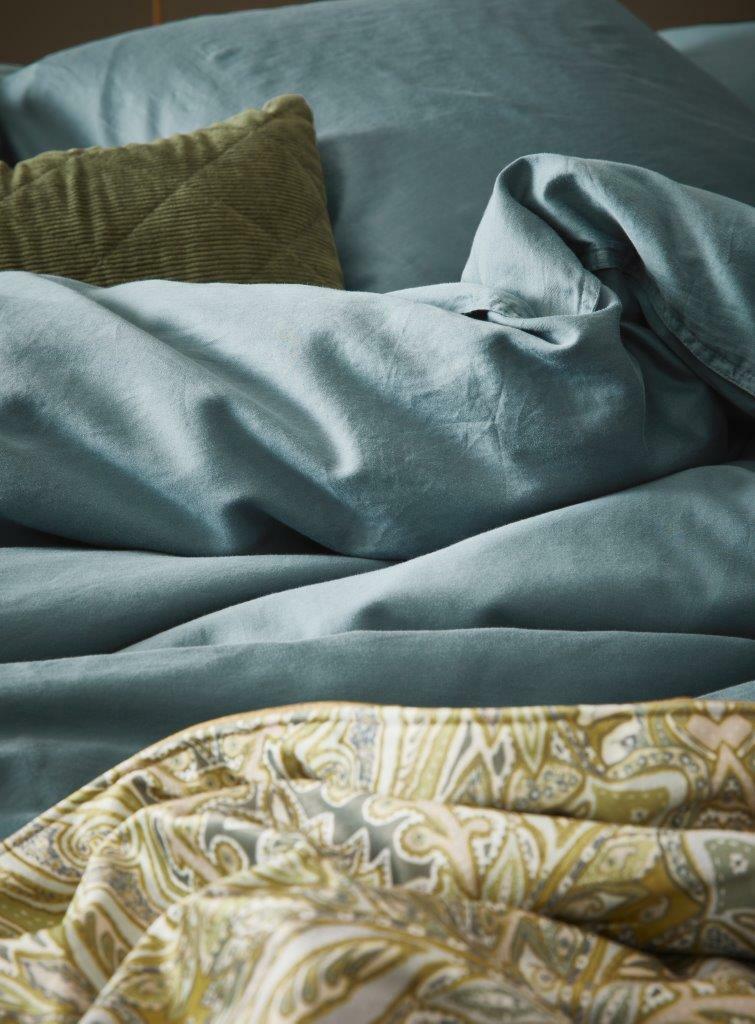 Nahaufnahme von Bettwäsche in sanftem Blau mit einem grünen Kissen und einem gemusterten Überwurf im Vordergrund.