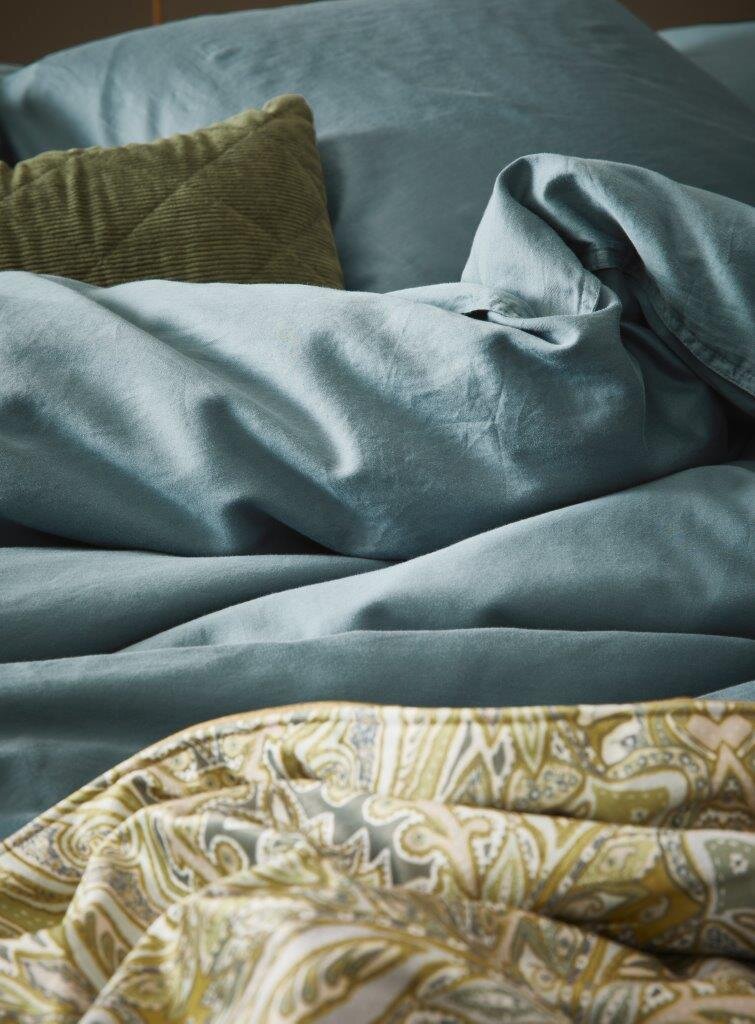Close-up van beddengoed in zachtblauw met een groen kussen en een gedessineerde sprei op de voorgrond.