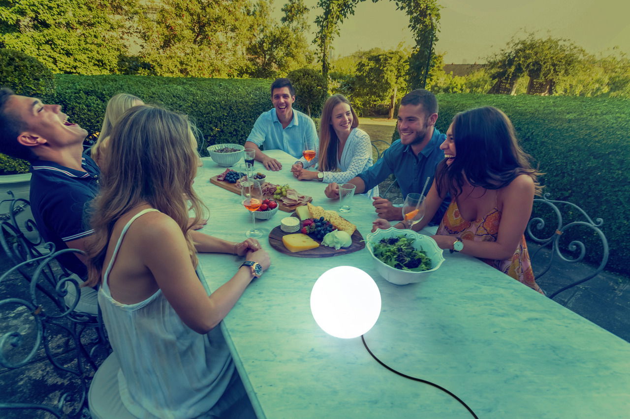 Kugelleuchte BULY 20 auf einem Tisch in einem Garten, umgeben von einer Gruppe lachender Menschen bei einem Abendessen, seitliche Perspektive.