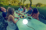 Kugelleuchte BULY 20 auf einem Tisch in einem Garten, umgeben von einer Gruppe lachender Menschen bei einem Abendessen, seitliche Perspektive.
