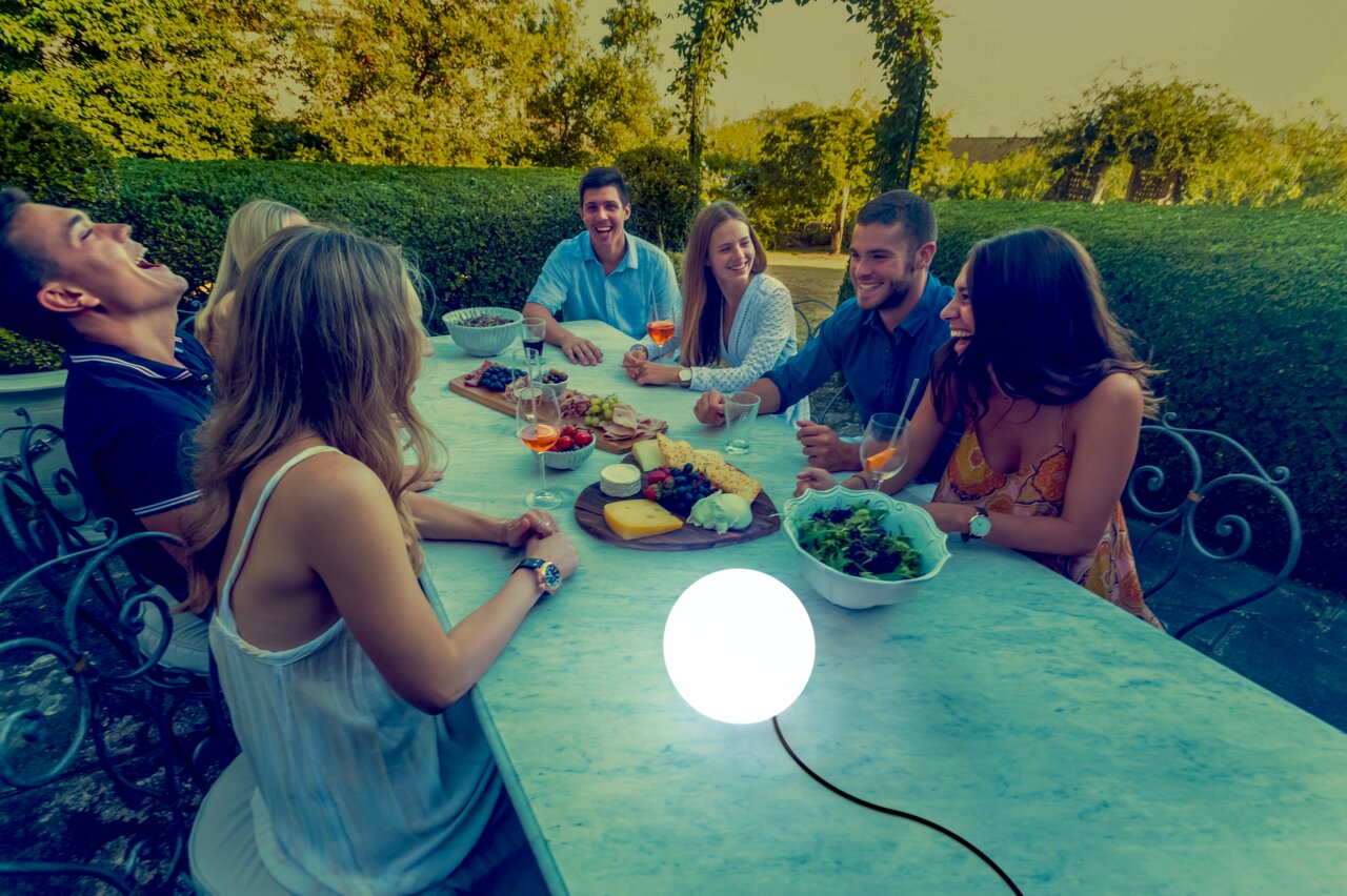 Kugelleuchte BULY 20 auf einem Tisch in einem Garten, umgeben von einer Gruppe lachender Menschen bei einem Abendessen, seitliche Perspektive.