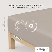 Spannbettlaken aus Bio-Baumwolle und Elasthan in Beige, auf einem Bett mit Holzrahmen, von der Seite betrachtet. Verpackung 100% recycelbar.
