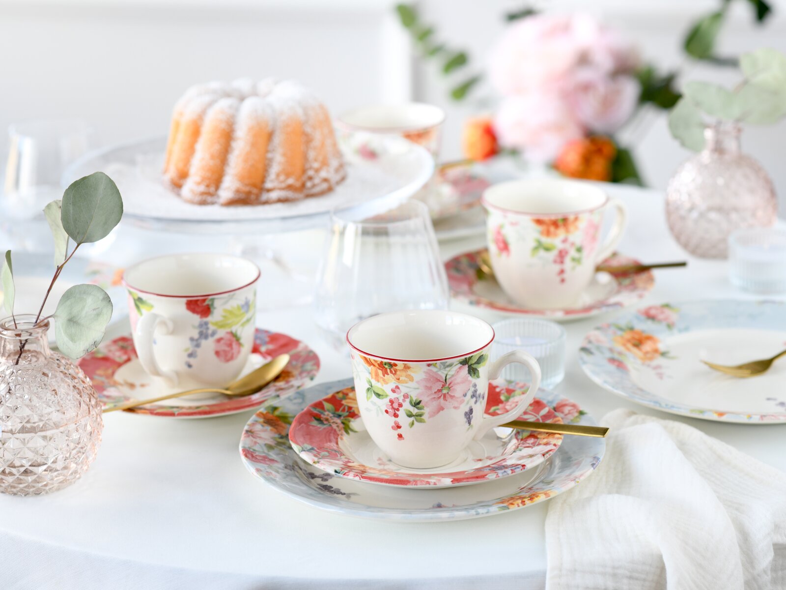 Florales Porzellan-Kaffeegeschirr mit Tassen, Untertassen und Tellern auf weiß gedecktem Tisch, im Hintergrund ein Gugelhupf – perspektivisch leicht von oben aufgenommen