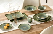 Grün-blau gesprenkeltes Keramikgeschirr mit Tellern, Schalen, Tasse und rechteckigen Platten, dekorativ auf einem Holztisch arrangiert, aus schräger Vogelperspektive.