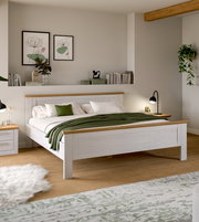 Modernes Doppelbett aus weißem Holz mit naturfarbenen Akzenten, weißer Bettwäsche und grüner Tagesdecke im hellen Schlafzimmer, perspektivisch in schräger Frontansicht von leicht rechts.