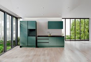 Moderne grüne Küchenzeile mit Schränken und Arbeitsplatte, frontal betrachtet, in einem hellen Raum mit großen Fenstern.
