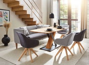 Moderner Esstisch mit Holzplatte und Metallgestell, umgeben von stilvollen Stühlen und einer Couch, in einem hellen Raum mit Treppe und großen Fenstern, seitliche Perspektive.