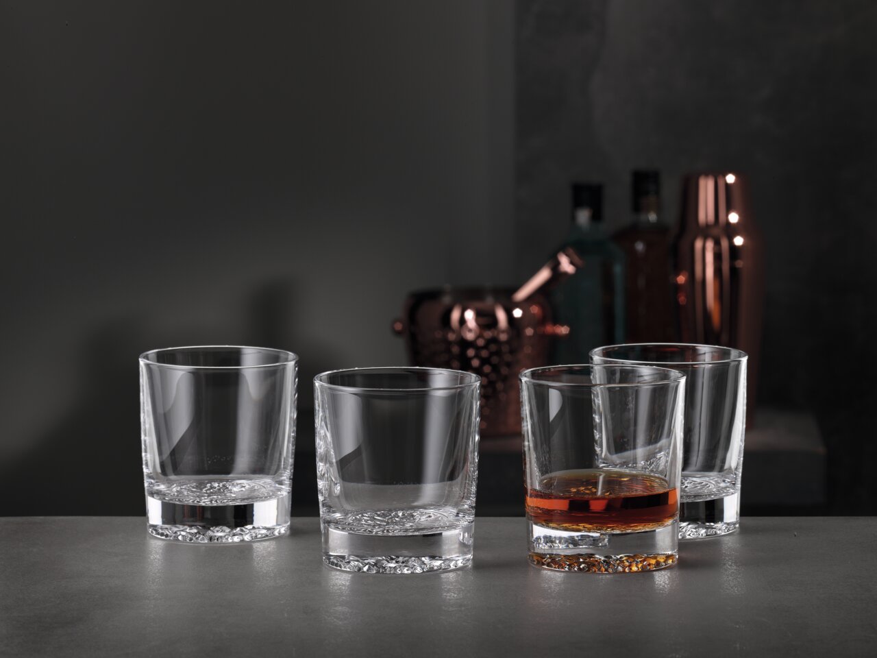 Spiegelau Whiskybecher 4er-Set LOUNGE Vier Whiskygläser in einem modernen, eleganten Design, eines davon mit Whisky gefüllt, auf einer grauen Oberfläche mit einer stilvollen Bar im Hintergrund, seitliche Perspektive.