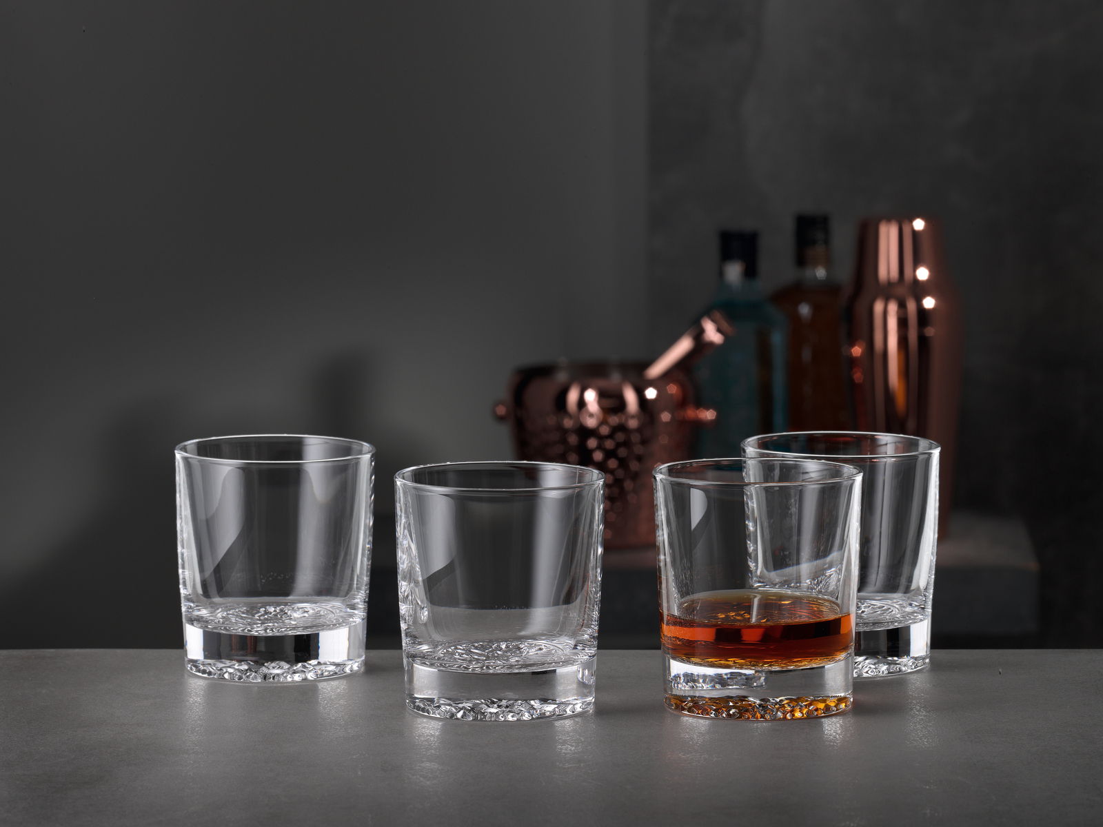 Vier Whiskygläser in einem modernen, eleganten Design, eines davon mit Whisky gefüllt, auf einer grauen Oberfläche mit einer stilvollen Bar im Hintergrund, seitliche Perspektive.