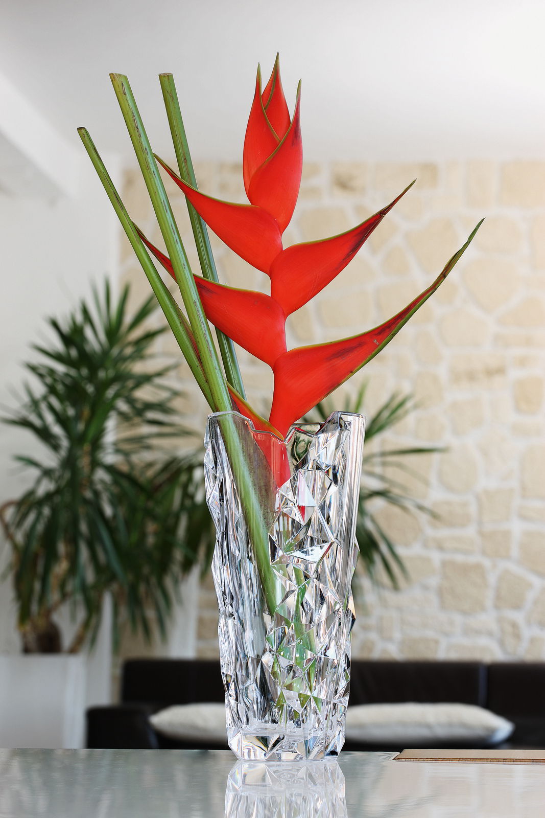 Kristallvase mit facettiertem Design, gefüllt mit exotischen roten Blumen, auf einem Tisch in einem modernen Wohnzimmer, seitliche Perspektive.