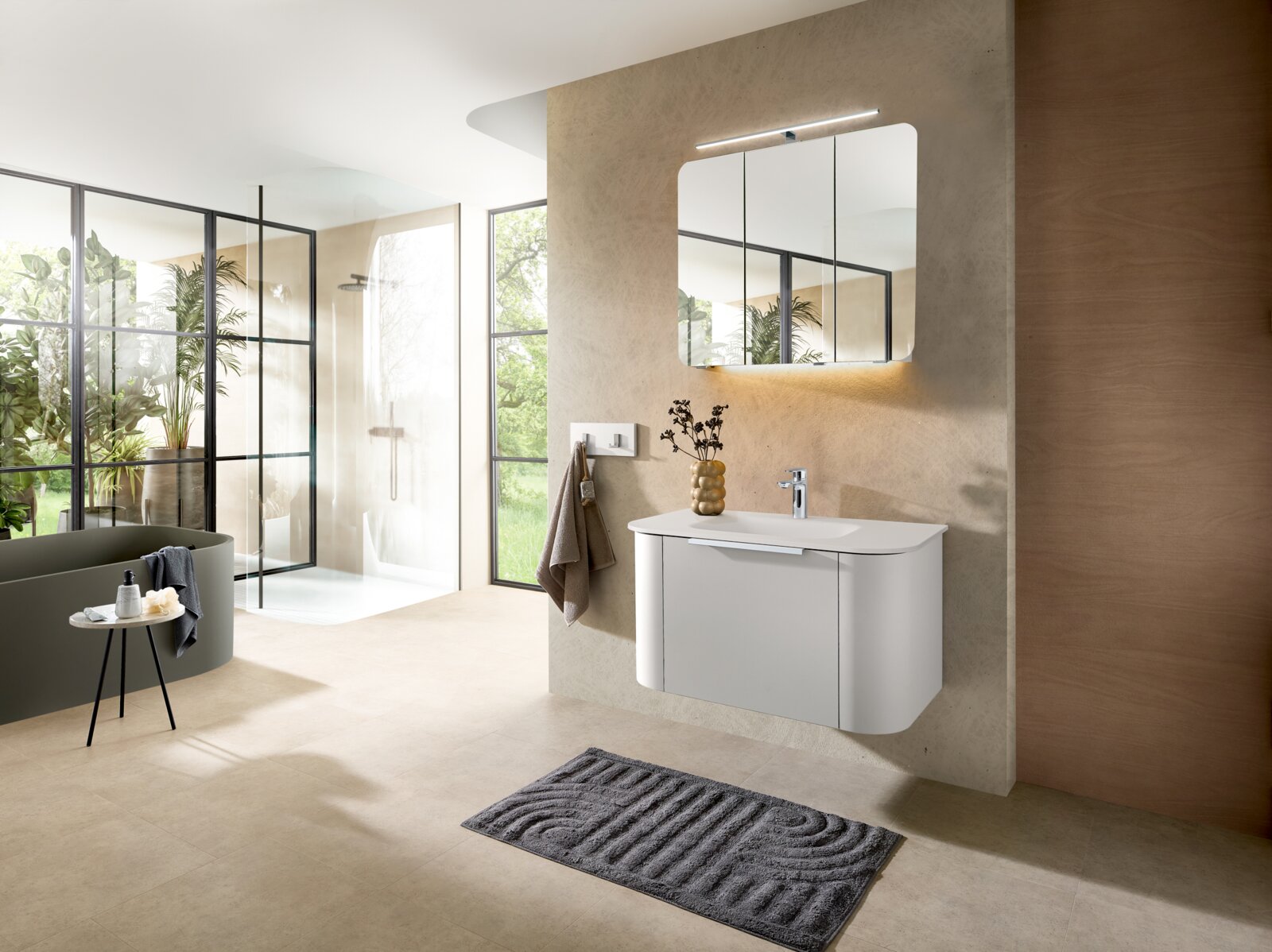 Modernes Badezimmer mit wandhängendem weißem Waschtischunterschrank, rechteckigem Spiegel mit LED-Leuchte und Einhebelmischer; Perspektive: schräger Blick von vorn rechts in den Raum mit Dusche und Badewanne im Hintergrund.