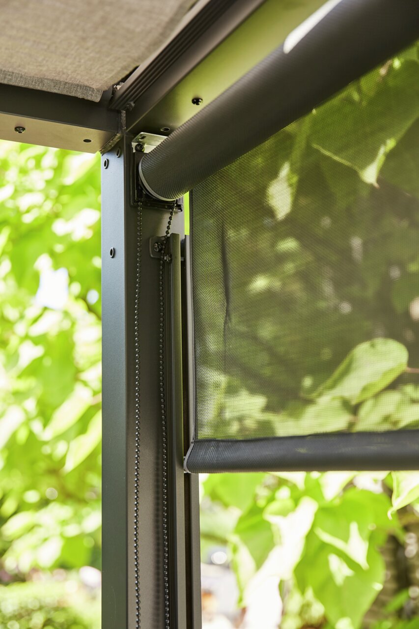 Close-up van een rolgordijn met kettingmechanisme, bevestigd aan een metalen frame, met groene bladeren op de achtergrond. Perspectief van onder naar boven.