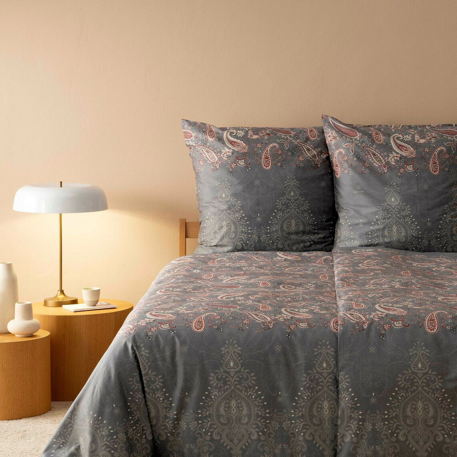 Bettwäsche-Set in Grau mit Paisley-Muster, bestehend aus einem Bettbezug und zwei Kissenbezügen, auf einem Bett mit Holzrahmen, seitliche Perspektive.