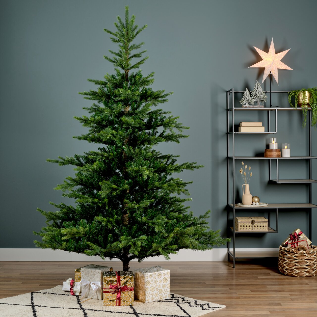 Künstlicher Weihnachtsbaum mit einer Höhe von 150 cm und einem Durchmesser von 111 cm, dekoriert mit Geschenken darunter, in einem modernen Wohnzimmer mit Regal und Dekorationen, seitliche Perspektive.