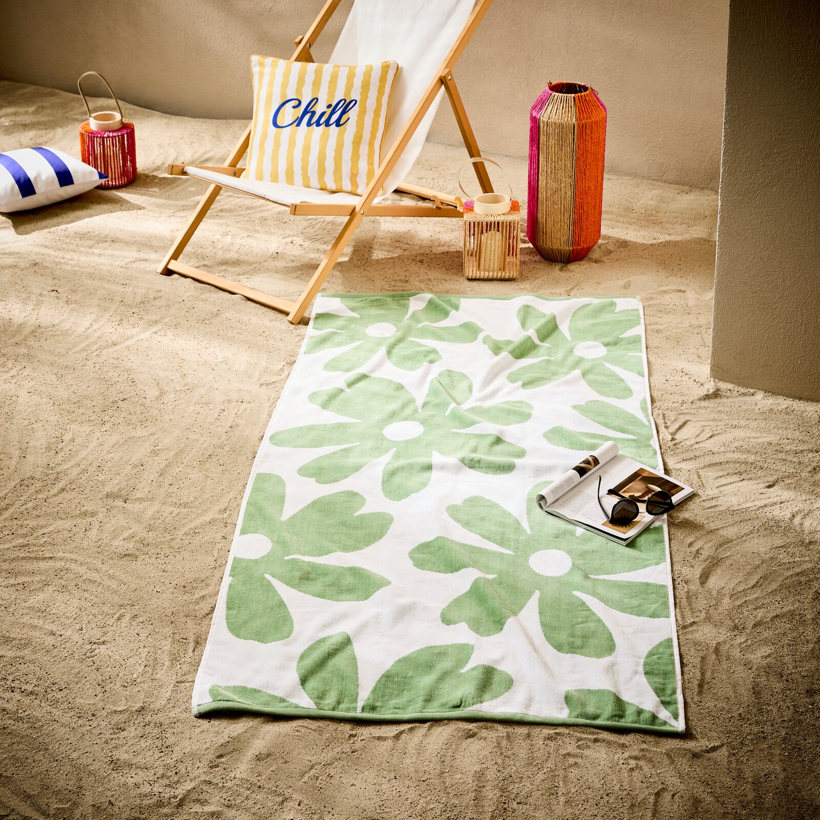 Esposa Strandtuch Schräg von oben fotografiertes Strandtuch in Weiß mit großem grünem Blumenmuster auf Sand, daneben eine Zeitschrift mit Sonnenbrille; im Hintergrund Liegestuhl mit Kissen und bunte Laternen.