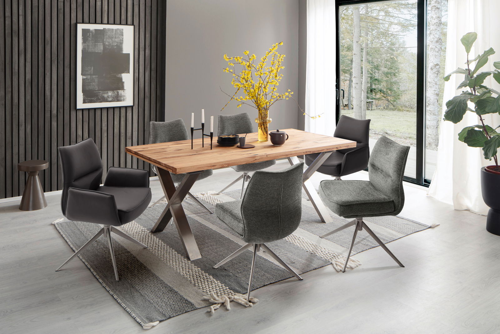 Esszimmer mit modernem Holztisch und sechs Stühlen, davon zwei mit Armlehnen. Die Stühle sind in Grau und Schwarz gehalten. Große Fenster im Hintergrund bieten einen Blick ins Freie.