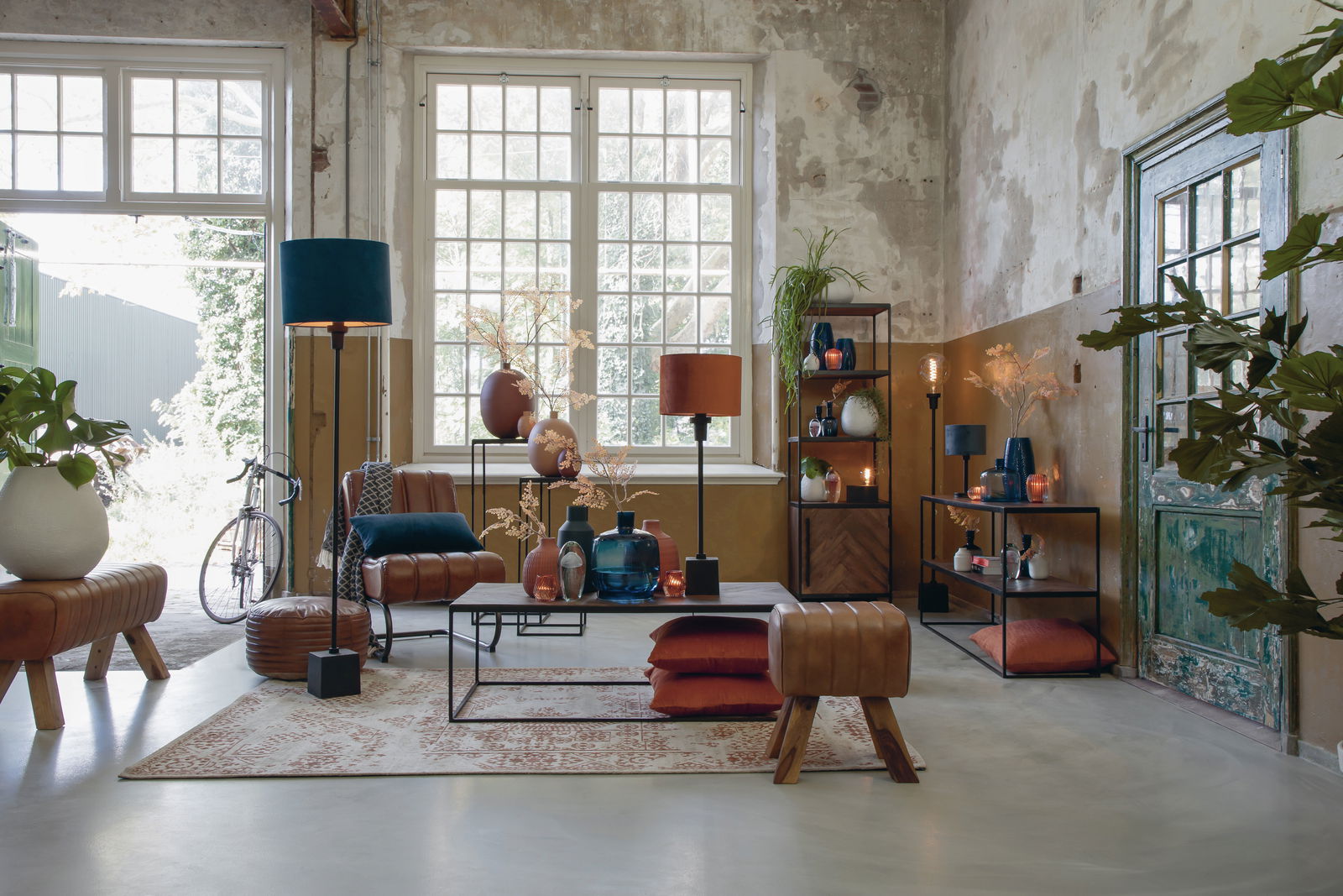 Industriell eingerichteter Raum mit großen Fenstern, einer braunen Lederbank im Vordergrund und verschiedenen Dekorationsgegenständen. Perspektive von vorne.