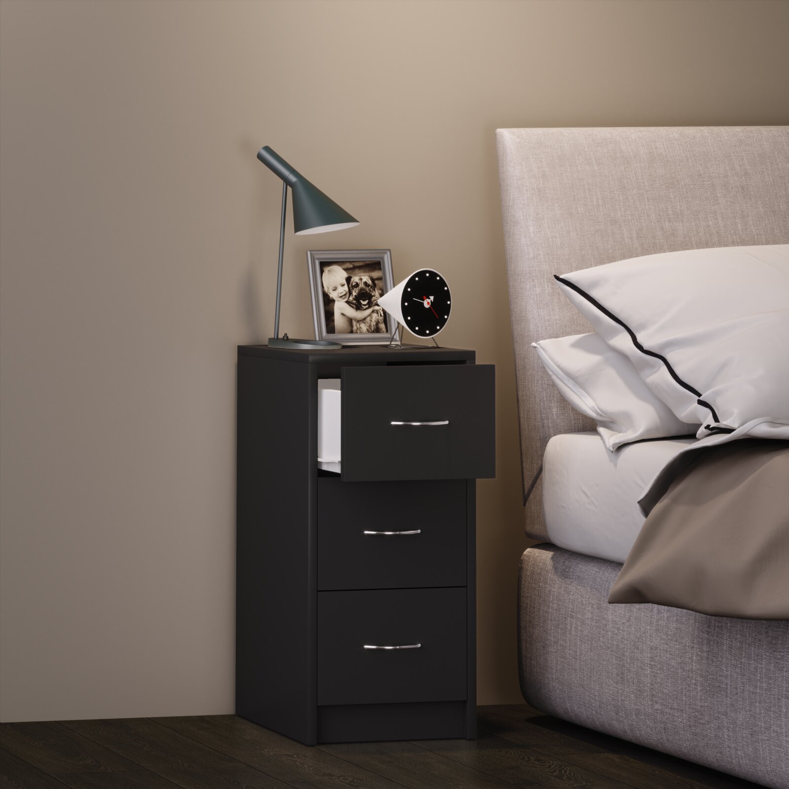 Schwarzer Nachttisch mit drei Schubladen neben einem gepolsterten Bett, darauf eine moderne Schreibtischlampe, ein Bilderrahmen und ein runder Wecker Ansicht schräg von vorn.