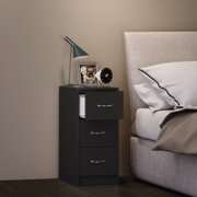 Schwarzer Nachttisch mit drei Schubladen neben einem gepolsterten Bett, darauf eine moderne Schreibtischlampe, ein Bilderrahmen und ein runder Wecker; Ansicht schräg von vorn.