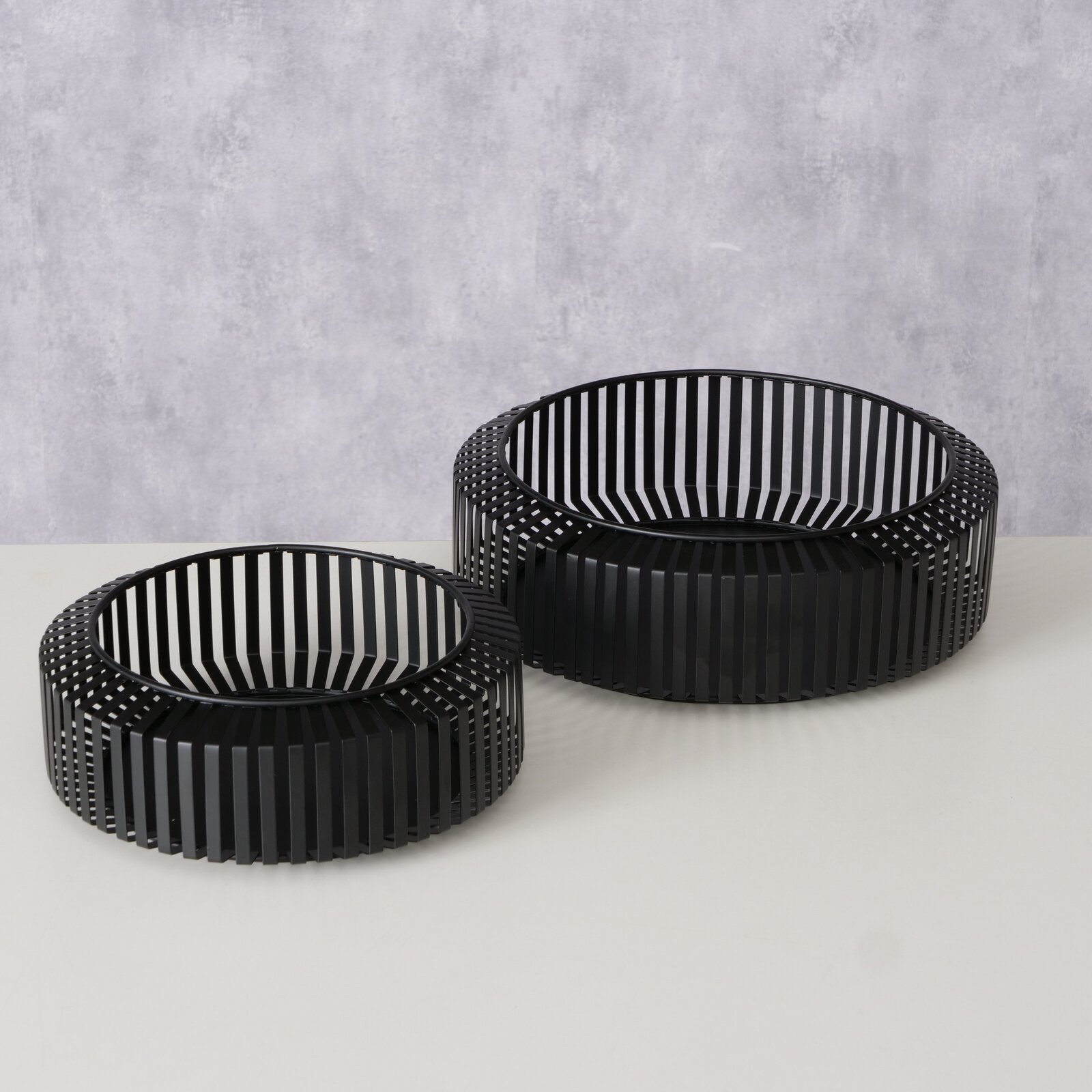 Zwei schwarze, runde Schalen mit vertikalen Streben, auf einer hellen Oberfläche stehend, aus einer leicht erhöhten Perspektive fotografiert.