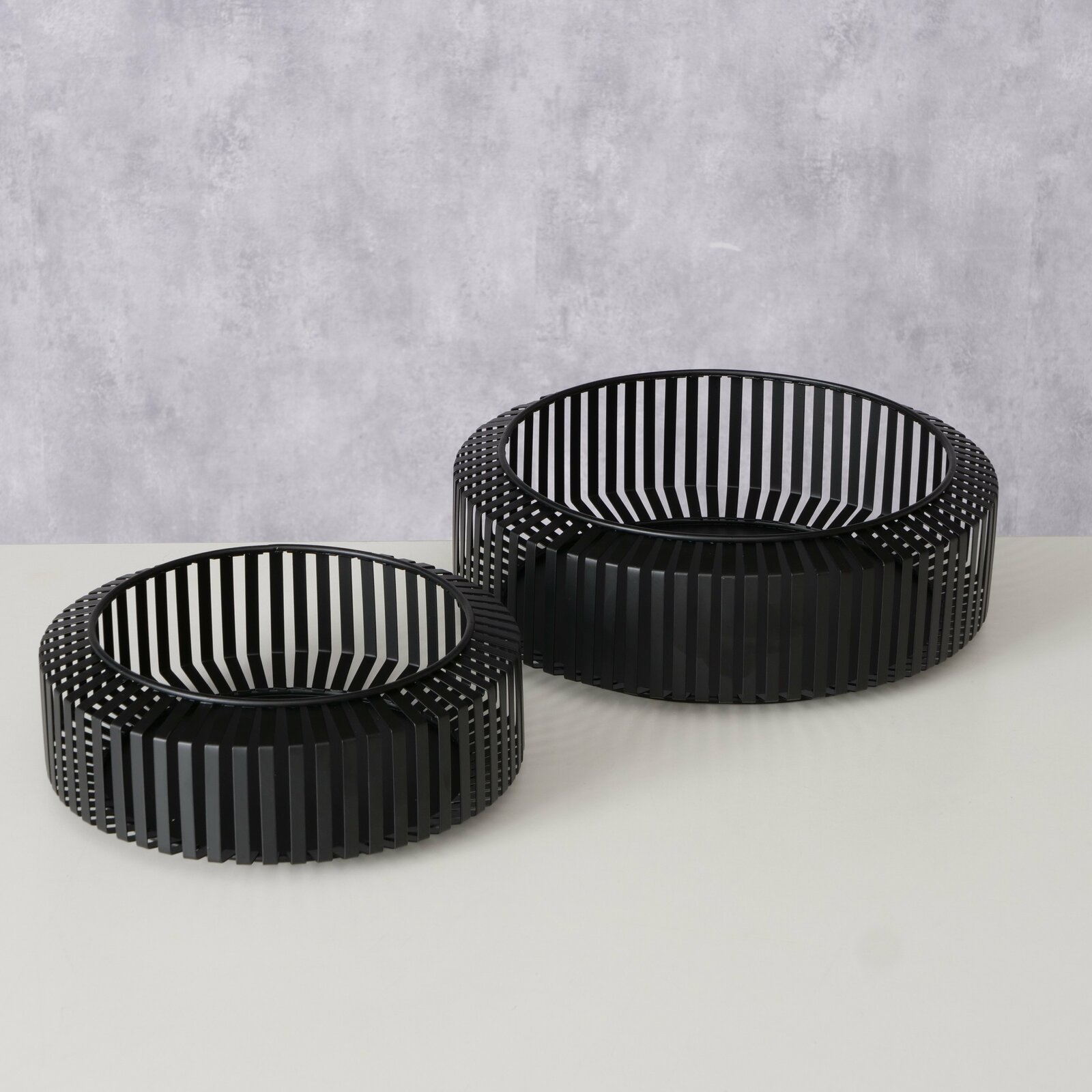 Zwei schwarze, runde Schalen mit vertikalen Streben, auf einer hellen Oberfläche stehend, aus einer leicht erhöhten Perspektive fotografiert.