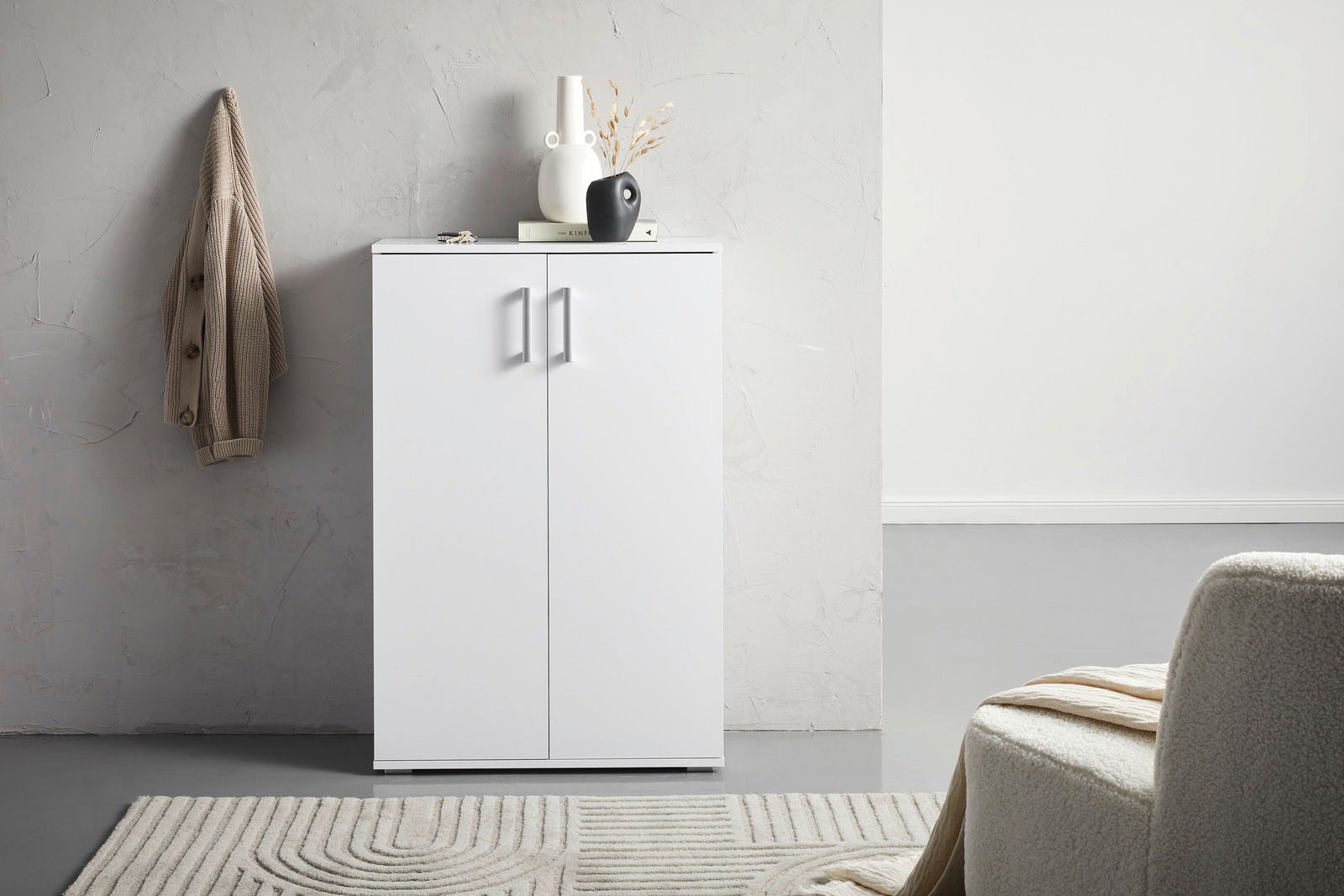 Moderner weißer Schuhschrank mit zwei Türen, frontal fotografiert, dekoriert mit Vasen und einem Buch, neben einer beigen Jacke an der Wand.