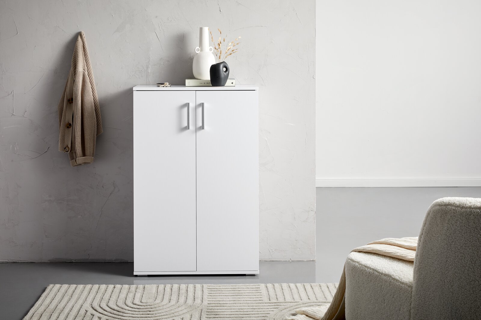 XORA Schuhschrank ALEX Moderner weißer Schuhschrank mit zwei Türen, frontal fotografiert, dekoriert mit Vasen und einem Buch, neben einer beigen Jacke an der Wand.