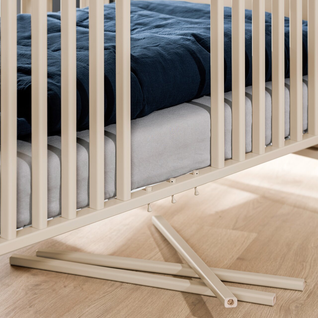 Close-up van een babybedje met blauw en grijs beddengoed, beige spijlen en een houten bodem, met twee verwijderde spijlen op de voorgrond.